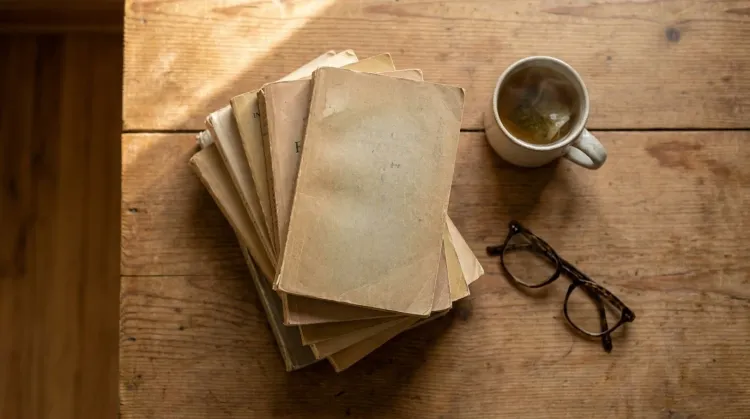 Overhead stack of well-worn paperback books beside a cup of tea and reading glasses on a wooden table