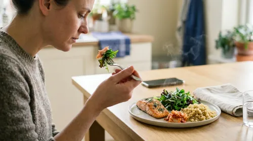 Person at a sunlit wood kitchen table mid-bite of mindful eating with salmon, greens, kimchi and quinoa on a pale plate