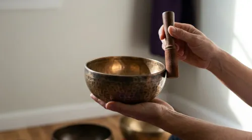 Practitioner's hands at a Tibetan singing bowl, wooden mallet poised at the rim, sound therapy in soft window-light