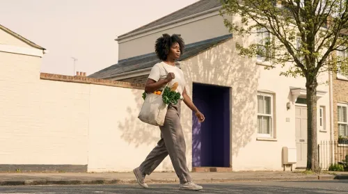 Woman walking home along a residential street with a canvas market bag of seasonal produce in soft late-morning light